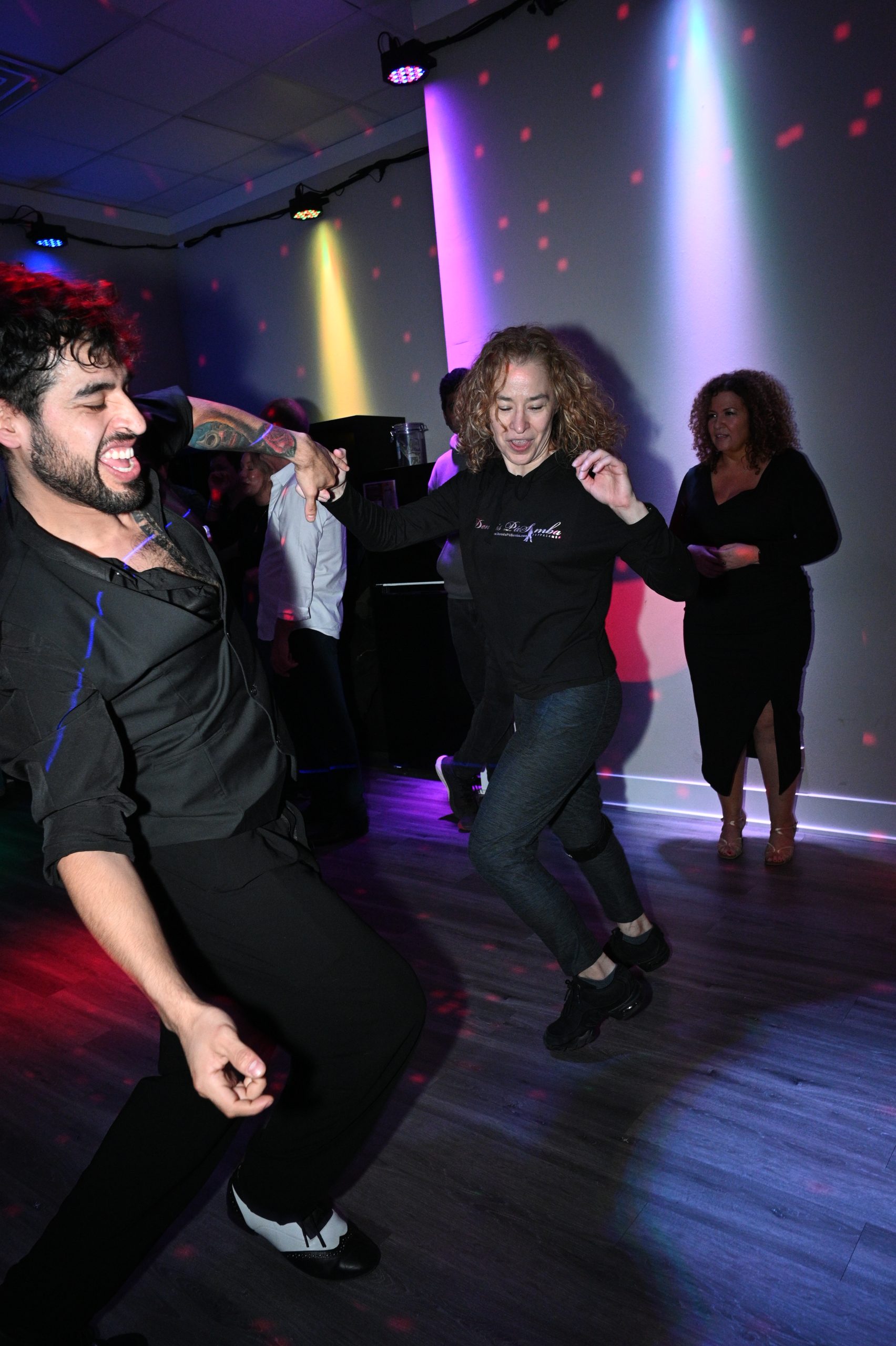 Two people are energetically dancing together in a dimly lit room with colorful spotlights, while others watch and smile in the background. The atmosphere appears lively and joyful.